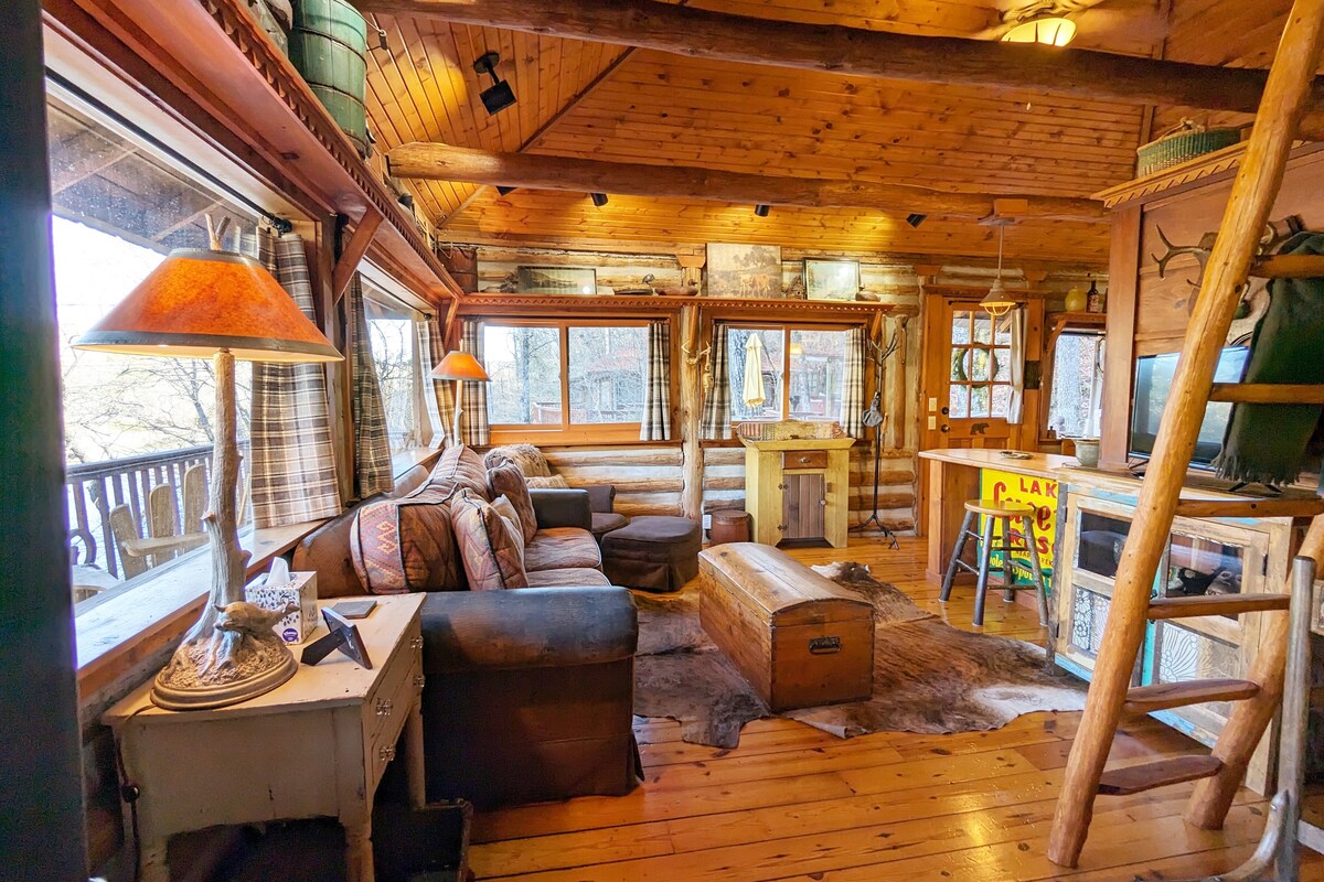 A cozy living area features wooden walls and ceiling beams, illuminated by warm light from a lamp. A comfortable sofa is arranged near a rustic coffee table, with a mix of decorative pillows. Windows offer views of the surrounding landscape, and a ladder leads to an upper level.