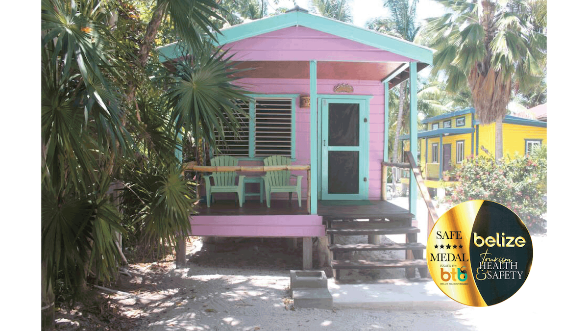 A vibrant cabana painted in shades of pink and turquoise is framed by lush greenery. A small porch area features a table and two green chairs, inviting guests to enjoy the tropical surroundings. Steps lead up to the entrance, enhancing accessibility.