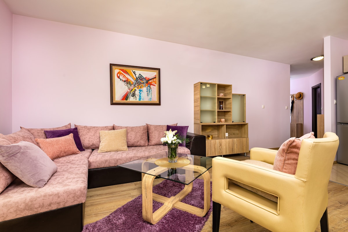 A cozy living area is furnished with a sectional sofa adorned with various cushions in soft hues. A glass coffee table sits at the center, complemented by a vase of fresh flowers. A colorful piece of art is displayed on the wall, adding a touch of personality.