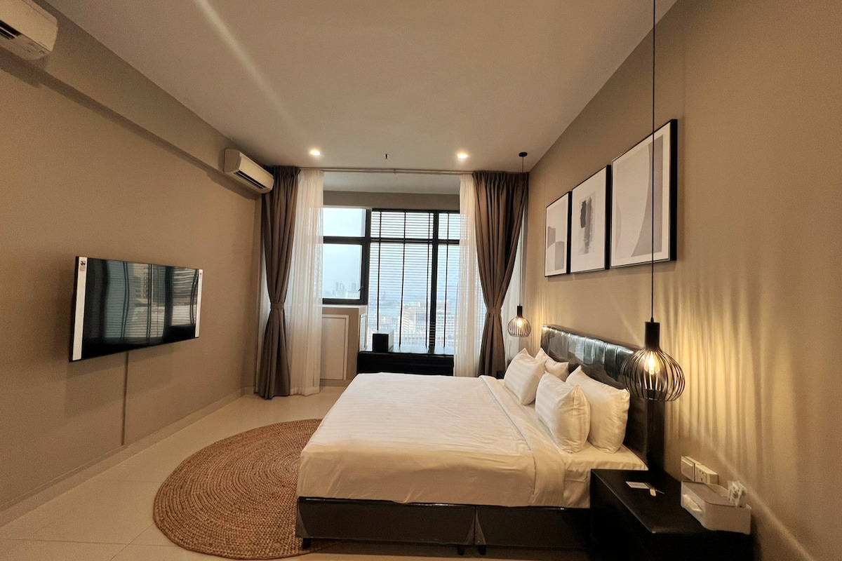 A spacious bedroom features a large bed with crisp white linens and decorative pillows. A flat-screen TV is mounted on the wall opposite large windows that offer natural light. Pendant lighting hangs above the bed, and a textured rug is positioned on the floor.