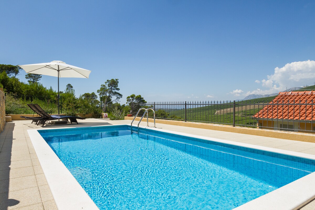 A heated pool is framed by a securely fenced area, featuring sun loungers and a large umbrella offering shade. The vibrant blue water reflects the clear sky, with gentle hills visible in the background providing a serene backdrop.
