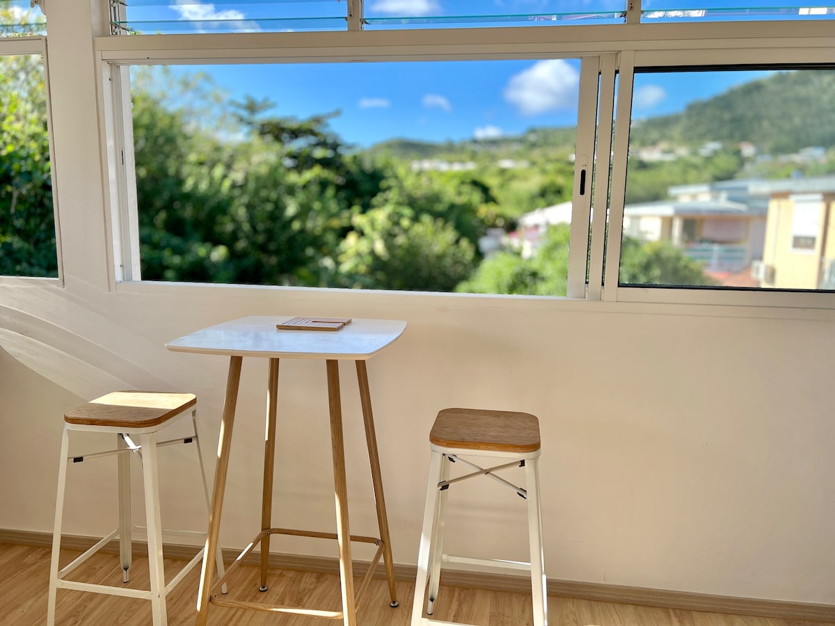 A bright dining area features a small table surrounded by two barstools. Large windows allow natural light to flood the space while offering views of greenery and rolling hills outside.