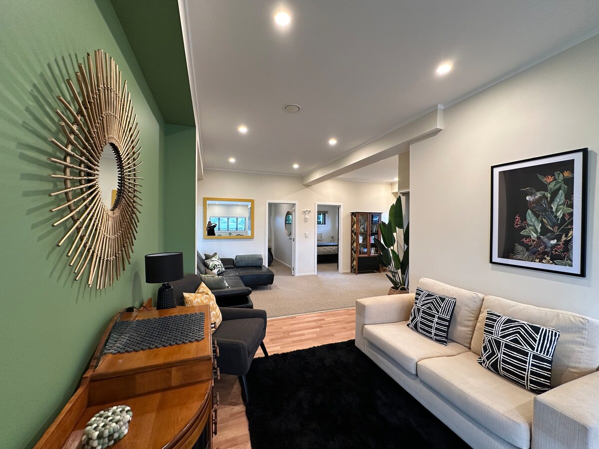 A spacious lounge area is featured, highlighted by a large mirror and carefully arranged decor. Comfortable seating options include a light sofa and dark armchairs. A rich green accent wall provides depth, while the natural light filters through large windows, enhancing the inviting atmosphere.