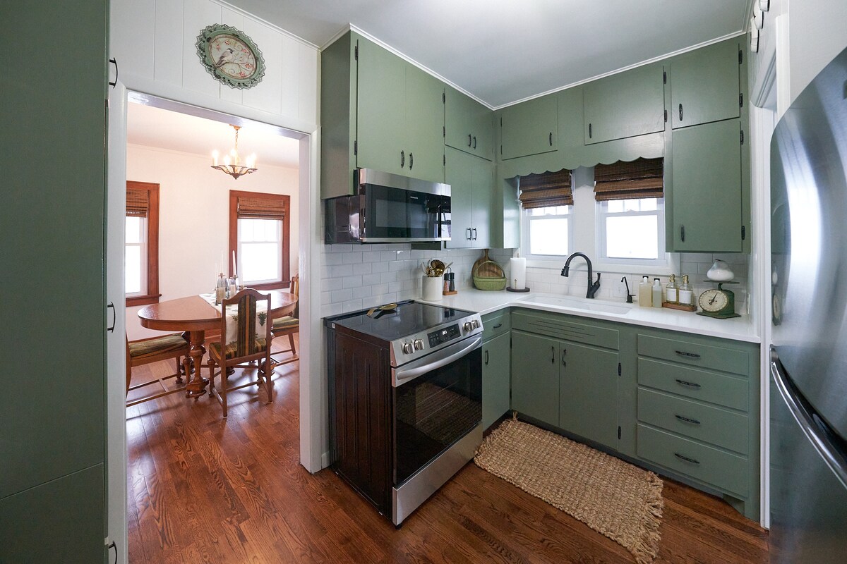 The kitchen features green cabinetry and modern appliances, including a stainless steel stove and microwave. A farmhouse-style sink is positioned beneath a window, with light-colored countertops offering a clean look. A view into the dining area reveals a wooden table surrounded by chairs.