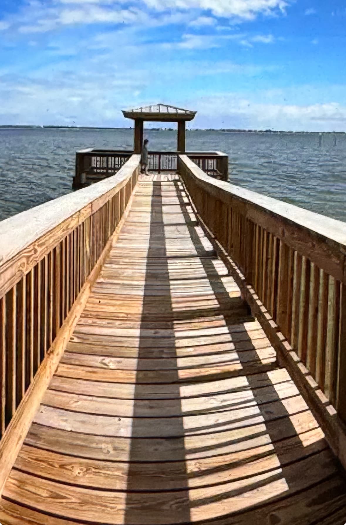 A wooden dock extends towards calm waters under a bright blue sky. Shadows from the railing create patterns along the walkway, leading to a shaded viewing area at the end. The scene captures the serenity of the waterfront and the natural light reflecting on the water.