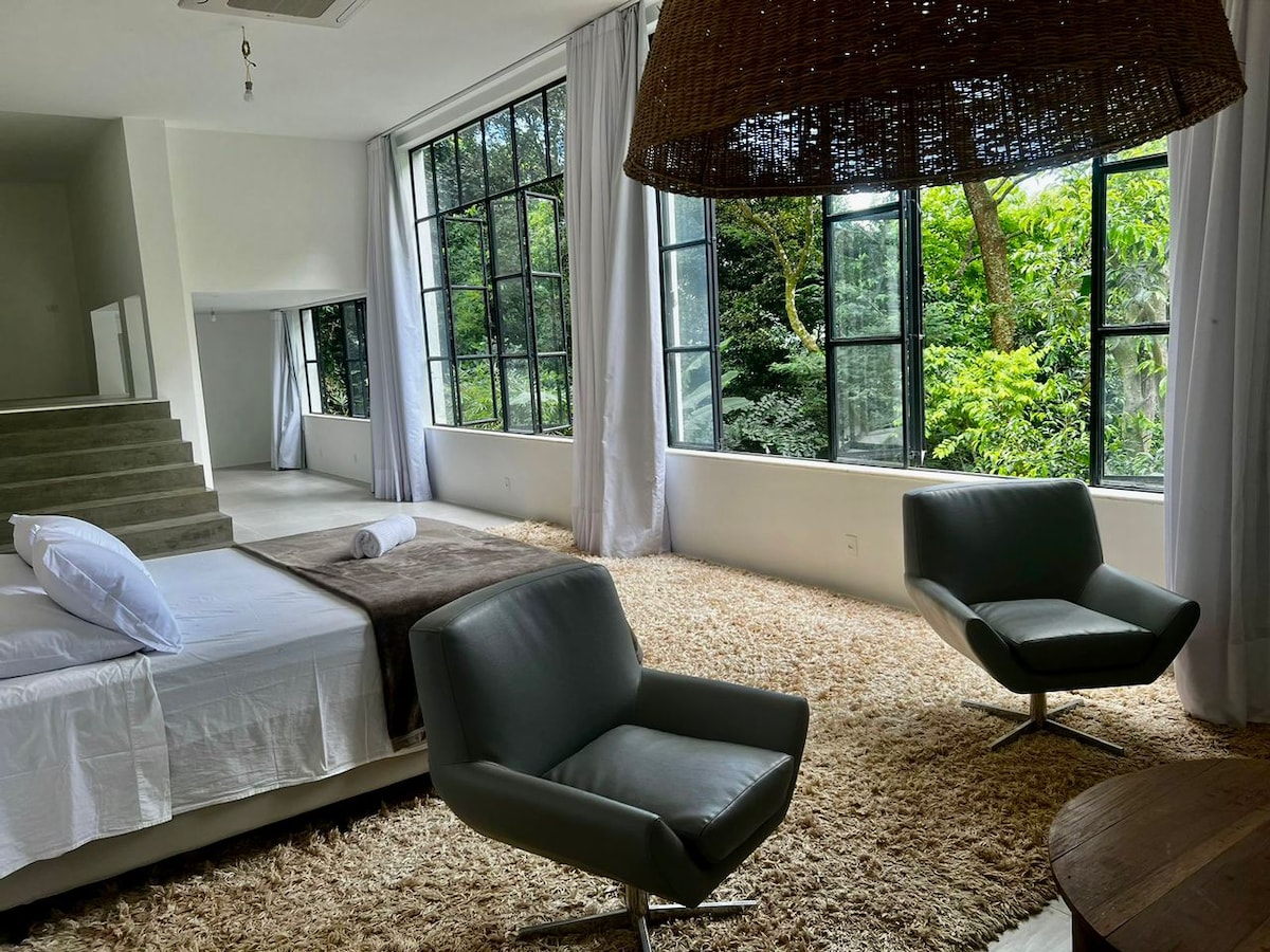 A spacious living area features large windows that invite natural light and offer views of the surrounding greenery. Two modern chairs are positioned on a textured rug, complemented by a cozy bed draped in neutral bedding, creating a harmonious connection with nature.