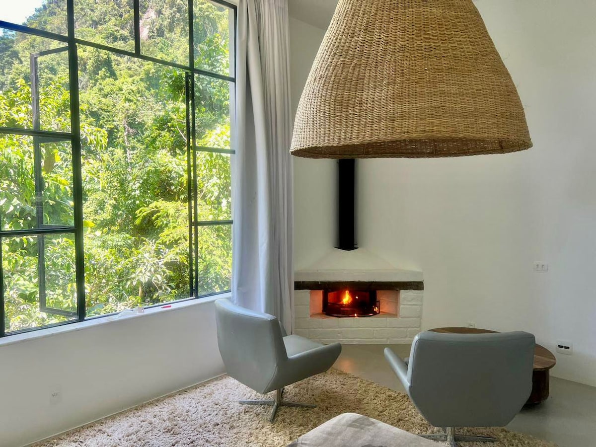 A cozy living area features a modern fireplace with a warm glow. Large windows open to a lush green landscape, allowing natural light to fill the space. Two sleek armchairs rest on a soft rug, creating a serene atmosphere for relaxation.