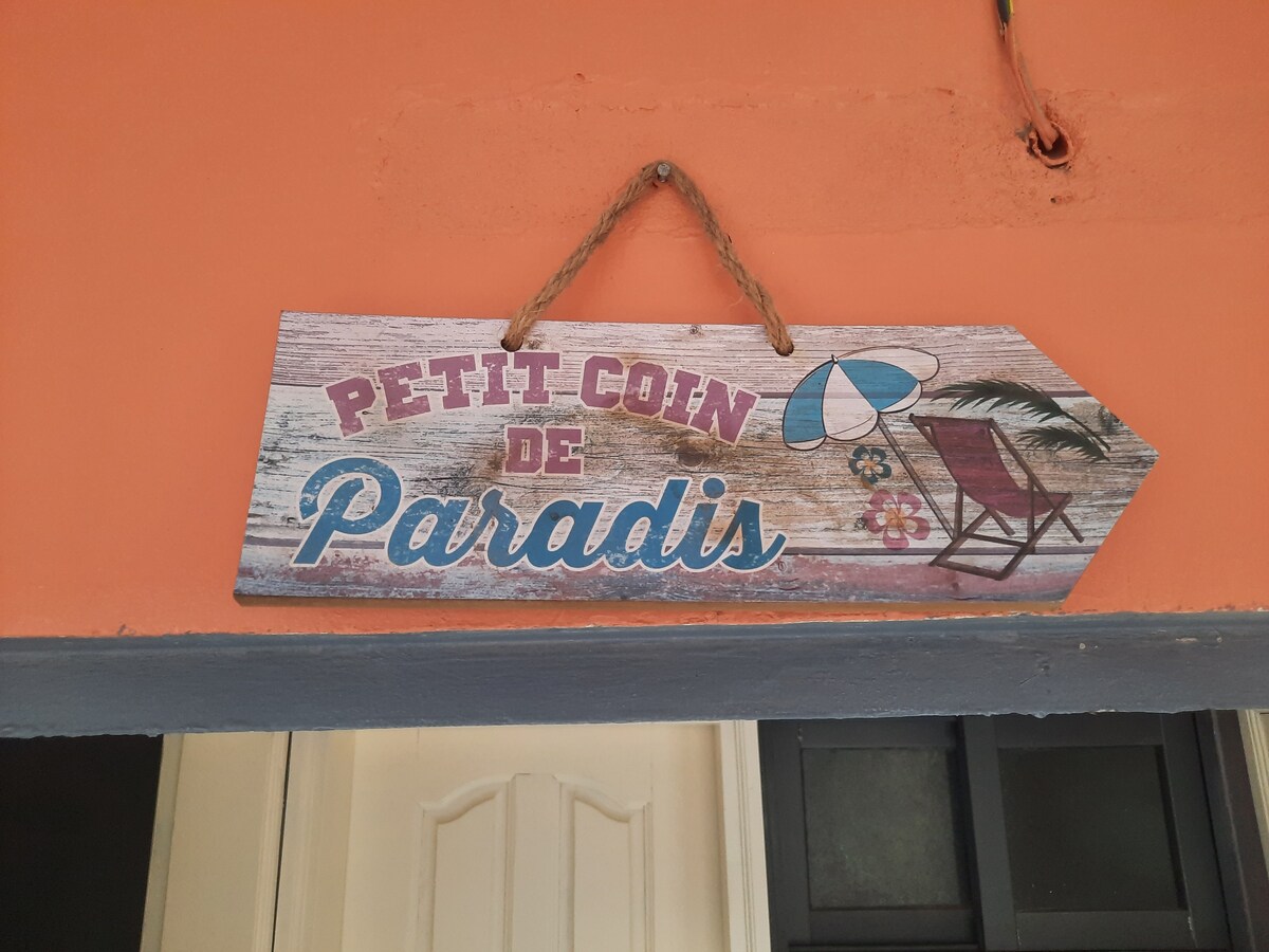 A wooden sign reading 'Petit Coin de Paradis' is displayed above a doorway. The sign features a coastal design, including an umbrella and a deck chair. The bright orange wall adds a cheerful touch to the entrance.