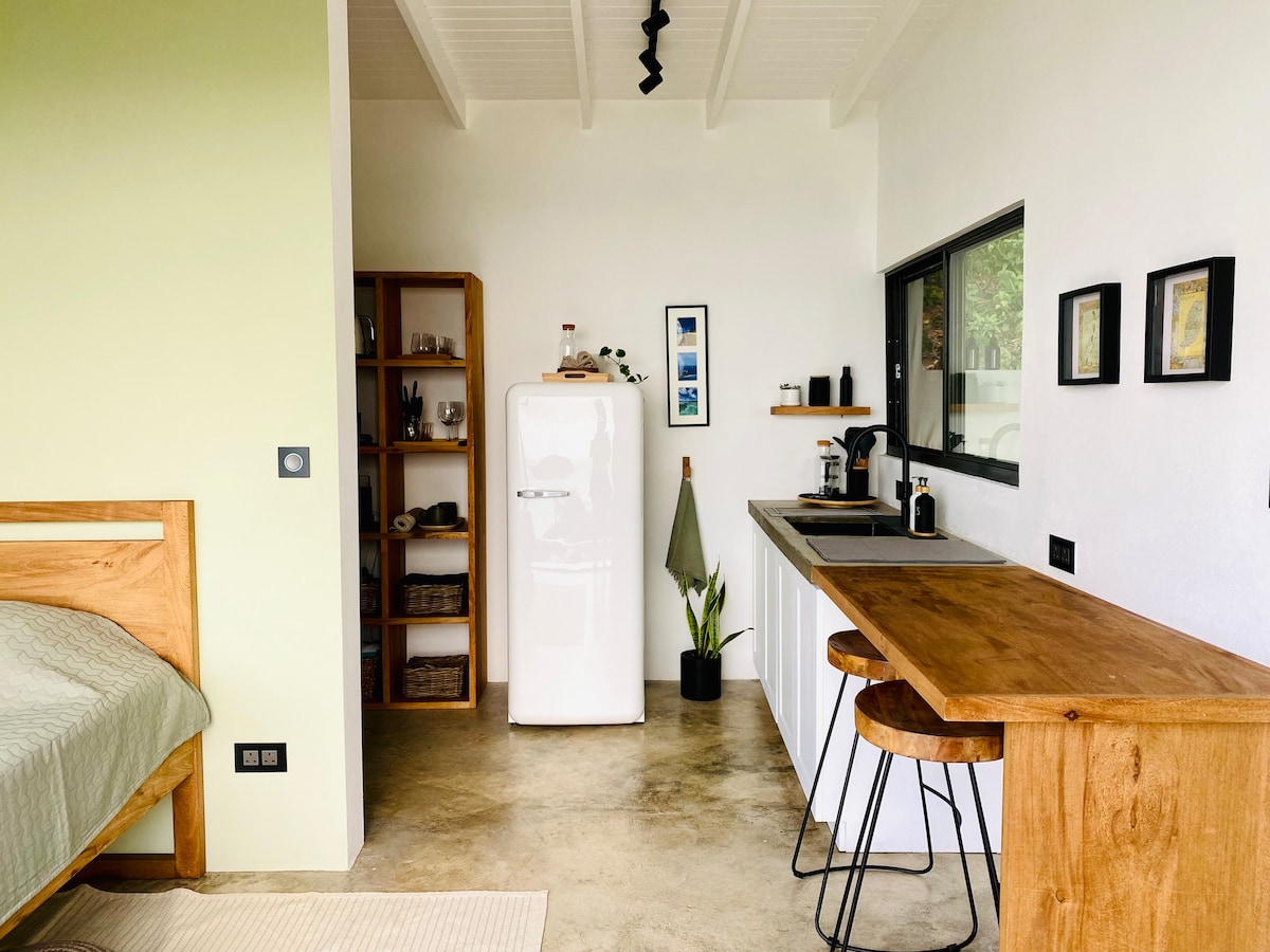 An open-concept interior features a modern kitchenette with a retro-style refrigerator and a wooden bar with two stools. Natural light filters through a large window, illuminating the minimalistic shelves and decor, creating a comfortable and inviting space.