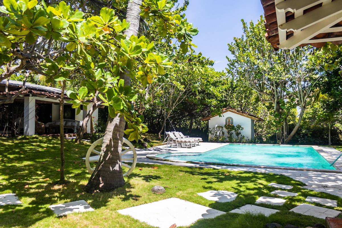 A serene outdoor area features a private pool bordered by lush greenery and white stone paths. Sun loungers are arranged near the pool, inviting relaxation. The surrounding trees provide shade, while a glimpse of a charming structure can be seen in the background.