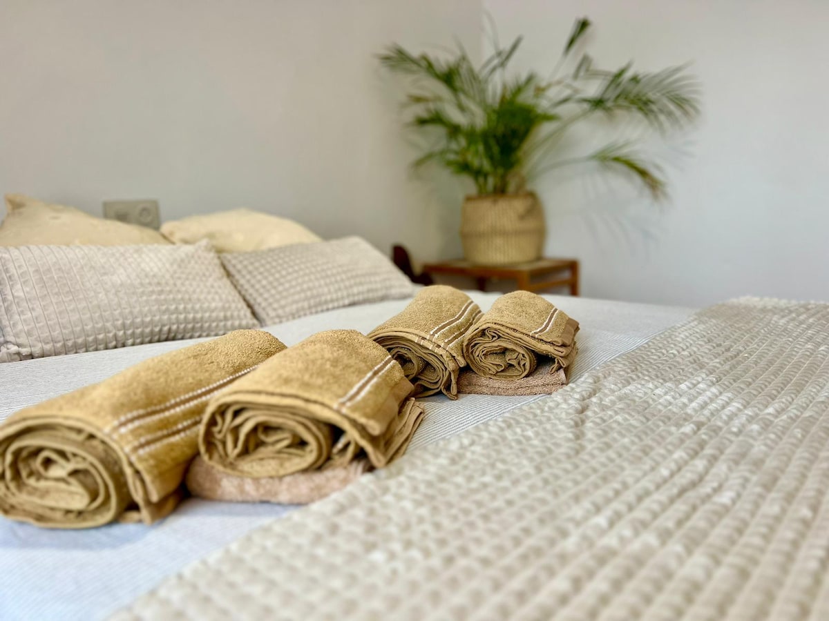 A spacious bed is presented with neatly folded golden towels. Textured cushions and a knitted throw add a soft touch. A plant in a woven pot enhances the calming environment, providing a sense of comfort and relaxation.