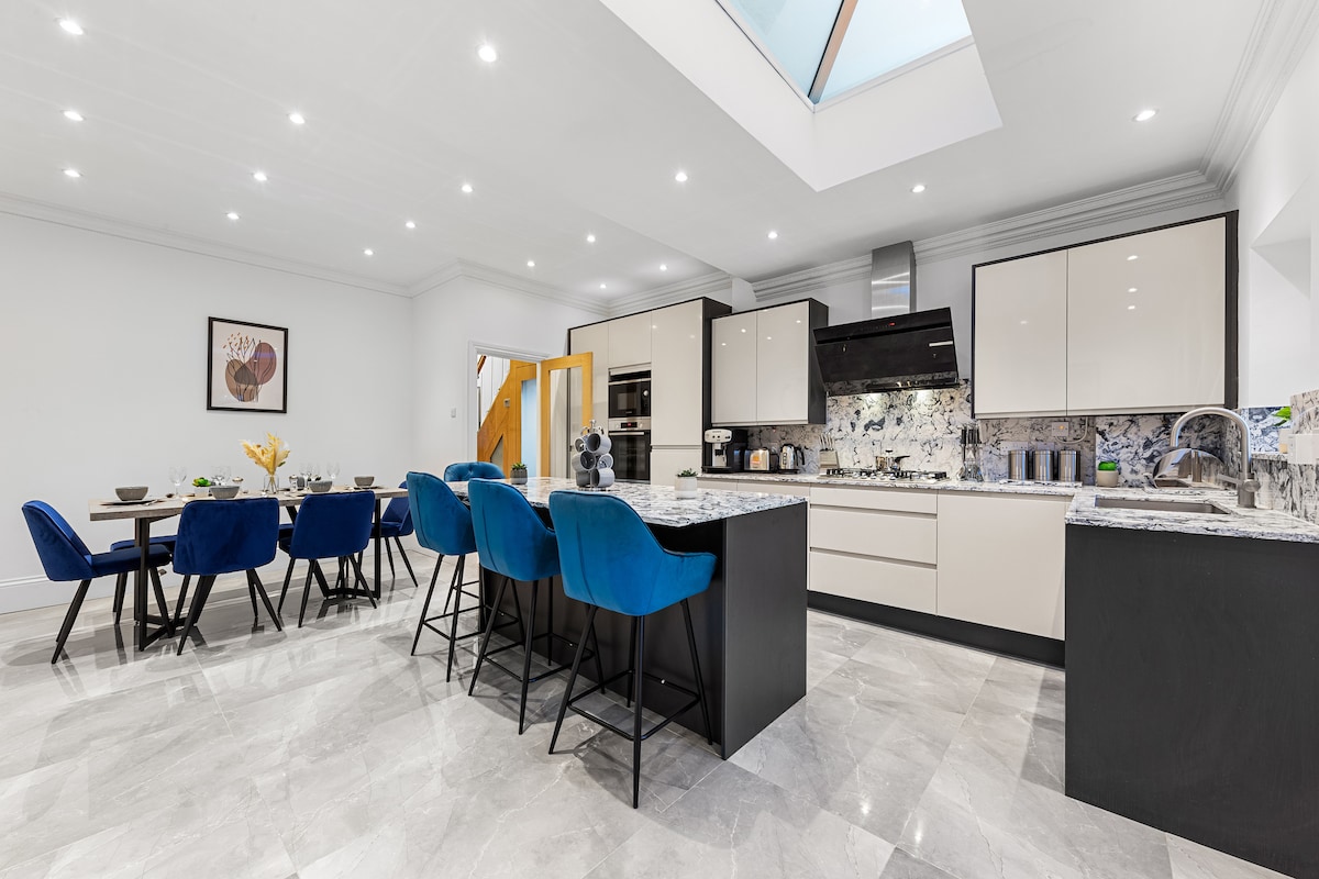 A modern kitchen features a spacious island with bar seating, complemented by a large dining table. Glossy cabinets and marble countertops create an elegant look, while ample natural light is provided through skylights. Stylish blue chairs enhance the contemporary aesthetic.