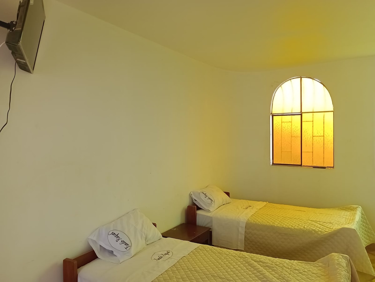 A well-lit room features two medium-sized beds neatly made with light-colored blankets. A wall-mounted television is visible, and a large arched window allows natural light to fill the space, framed by a grid of orange-tinted glass.