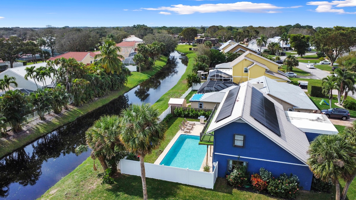 Aerial view of a waterfront property showcasing a backyard pool surrounded by palm trees. A canal runs alongside the property, while neighboring houses are visible in the distance. The bright blue exterior of the house contrasts with the lush greenery and vibrant sky.