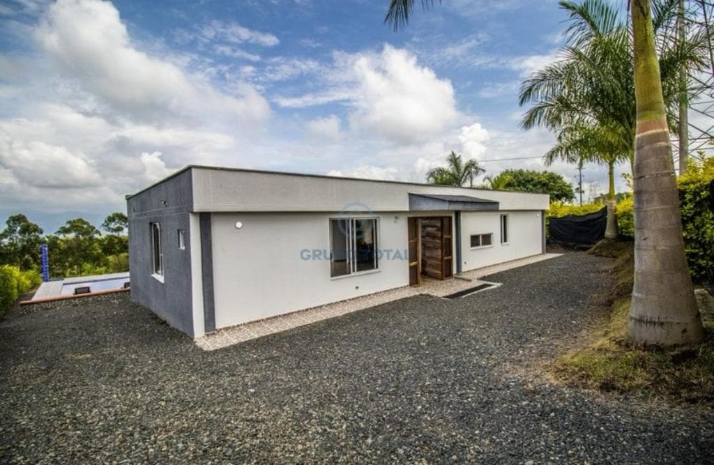 A modern single-story house is surrounded by gravel landscaping and palm trees. Large windows allow natural light to fill the living space, and a wooden door serves as a welcoming entrance. The peaceful setting is enhanced by a clear sky with scattered clouds.