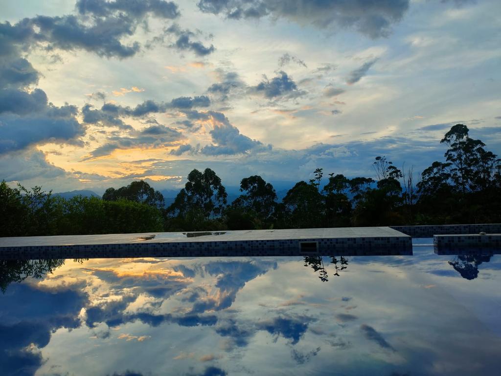 A calm pool reflects the vibrant hues of the sunset sky, surrounded by lush greenery and distant mountains. The serene landscape creates a peaceful aesthetic, presenting an inviting space for relaxation.
