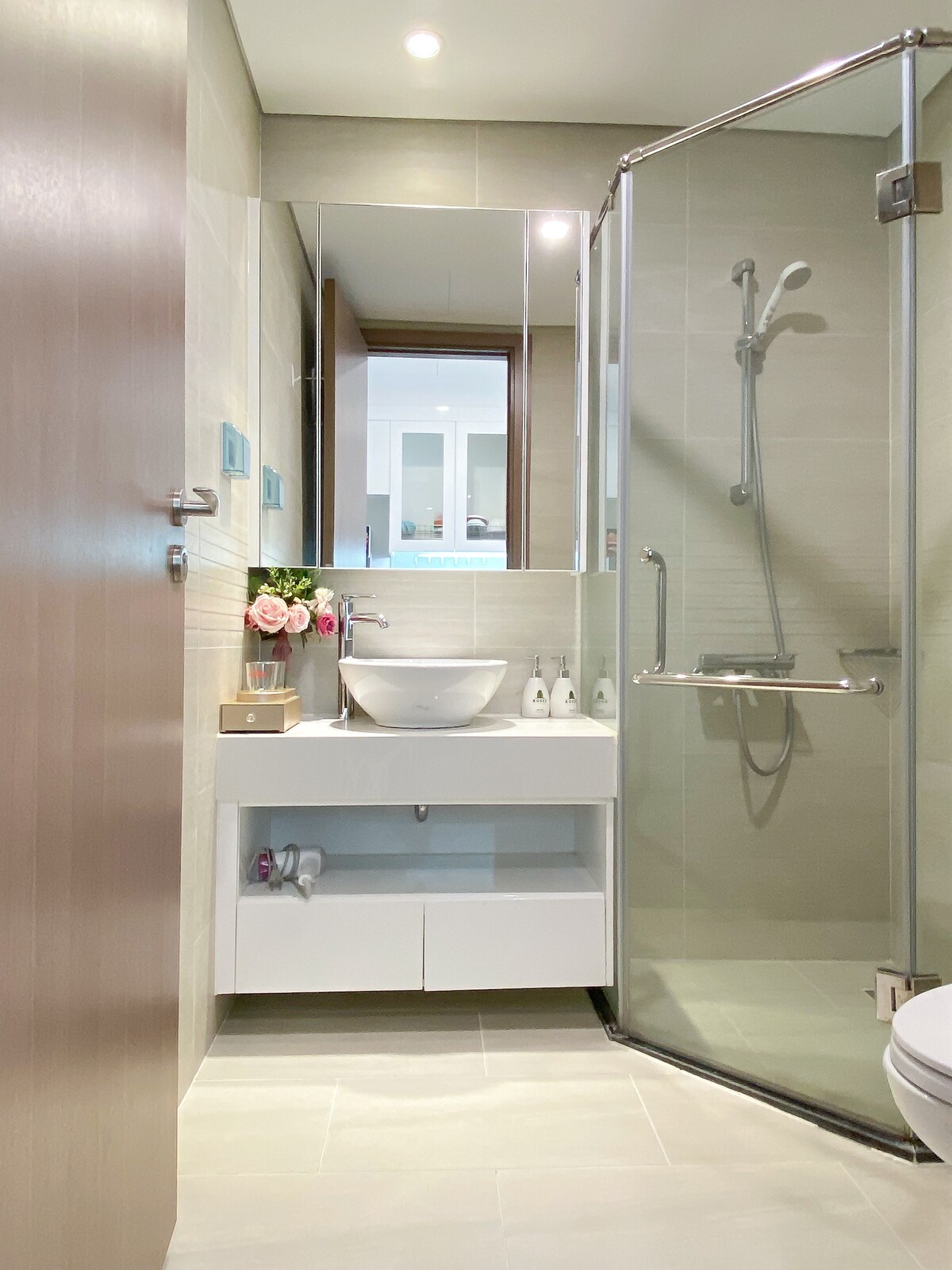 The bathroom features a modern design with a glass shower enclosure and a sleek white sink atop a minimalistic vanity. Soft neutral tones are present in the tiles and wall, complemented by a small vase of flowers, creating a fresh and serene atmosphere.