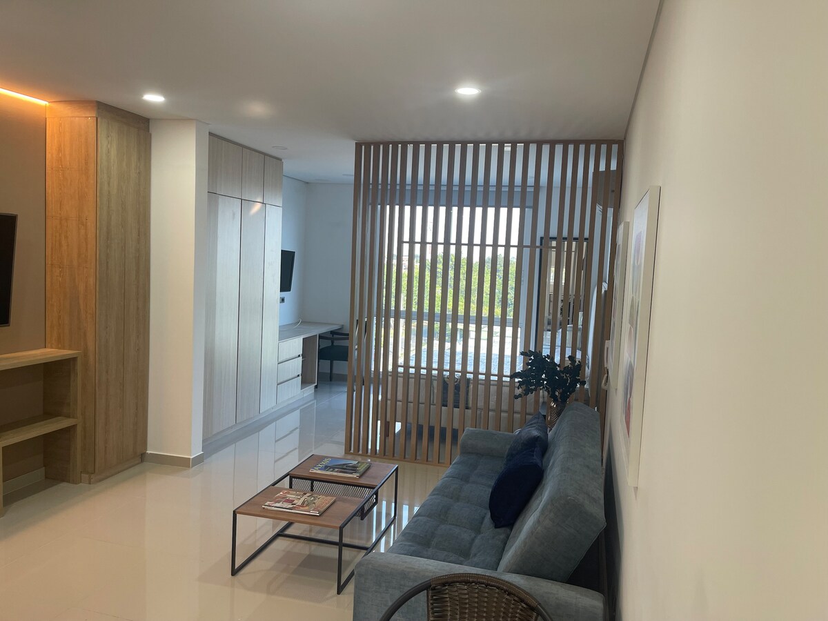 A modern living area features a comfortable sofa and a coffee table with magazines. A wooden partition adds texture to the space, separating the living area from the sleeping quarters bathed in natural light from the large window.