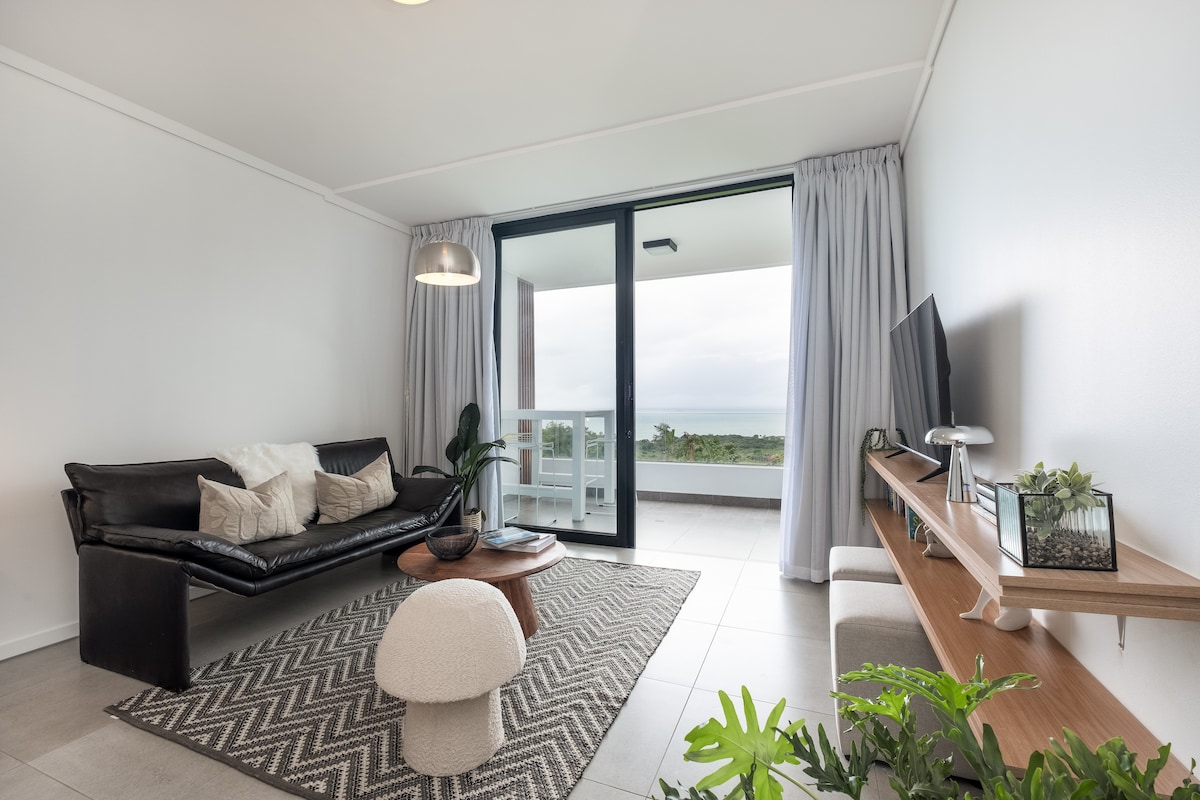 A modern living area features a plush black leather sofa, complemented by neutral-toned cushions. A round wooden table sits in front, atop a woven area rug. Large glass doors open to a balcony, with natural light enhancing the space. A minimalist wooden shelf holds decorative items.