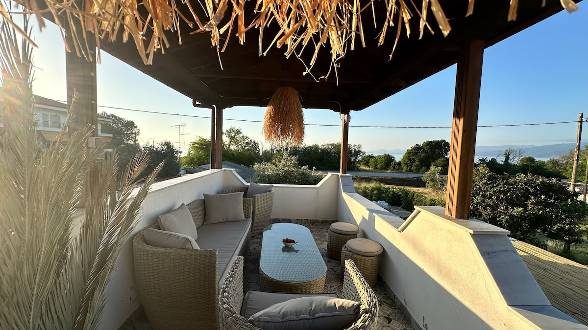 An outdoor chill-out zone is presented under a wooden pergola, featuring comfortable seating, a glass-top table, and round stools. Natural light filters through, highlighting the tranquil surroundings with greenery and distant hills visible in the background.