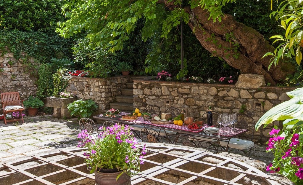 A stone courtyard is surrounded by lush greenery and blooming flowers. A long table is set for dining, displaying an array of fruits and drinks. Wooden chairs are positioned nearby, and a large tree provides shade, enhancing the inviting outdoor space.