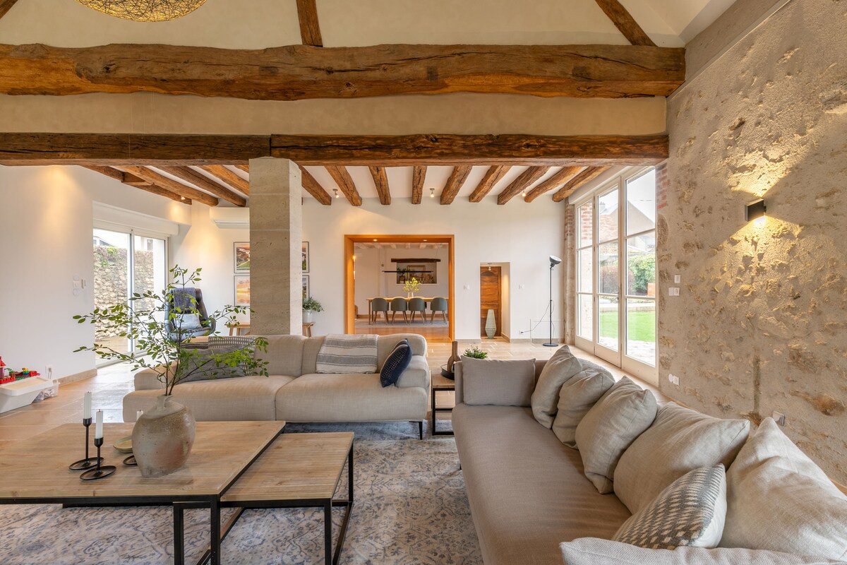 A spacious living area is highlighted by exposed wooden beams and natural light streaming through large windows. Comfortable sofas are arranged around a coffee table, creating an inviting space. The adjoining dining area is visible, featuring a long table surrounded by chairs.