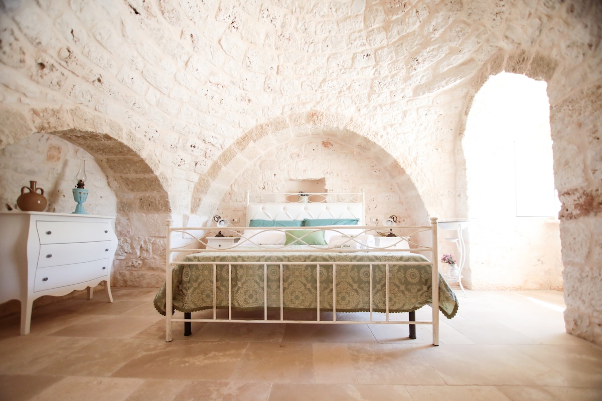 A spacious sleeping area features a metal bed frame with a patterned bedspread. Soft light enters from a nearby window, highlighting the stone arches that create a unique architectural detail. A white dresser is situated beside the bed.