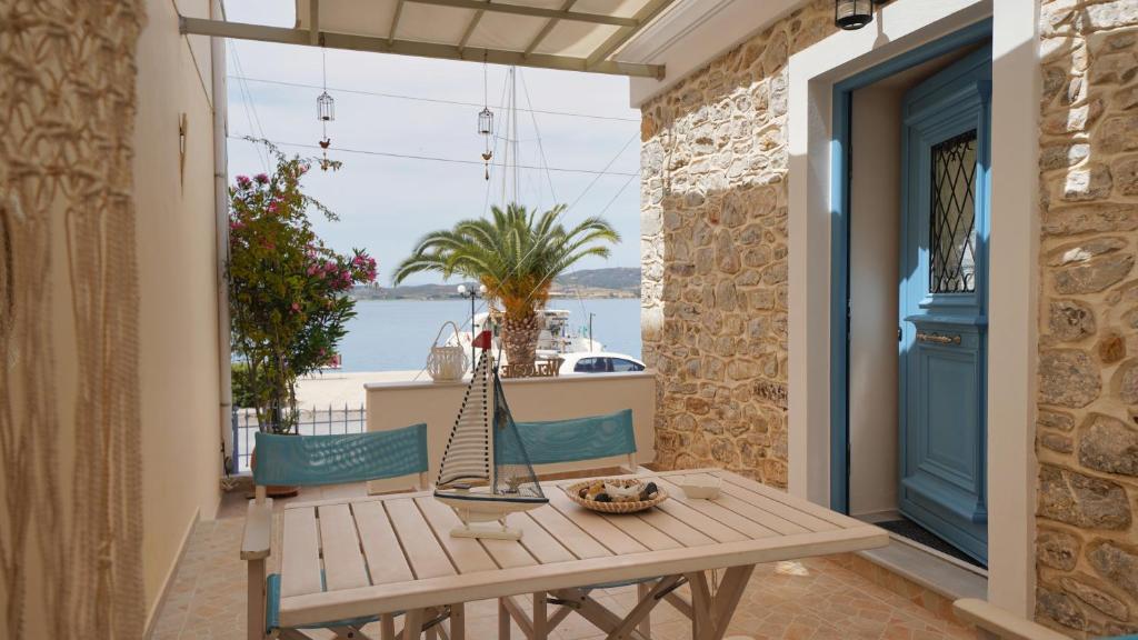 A shaded patio area features a wooden table surrounded by blue chairs. A decorative sailboat centerpiece and a bowl of snacks sit on the table. The stone wall adds texture, while a view of the sea and palm trees is visible in the background.