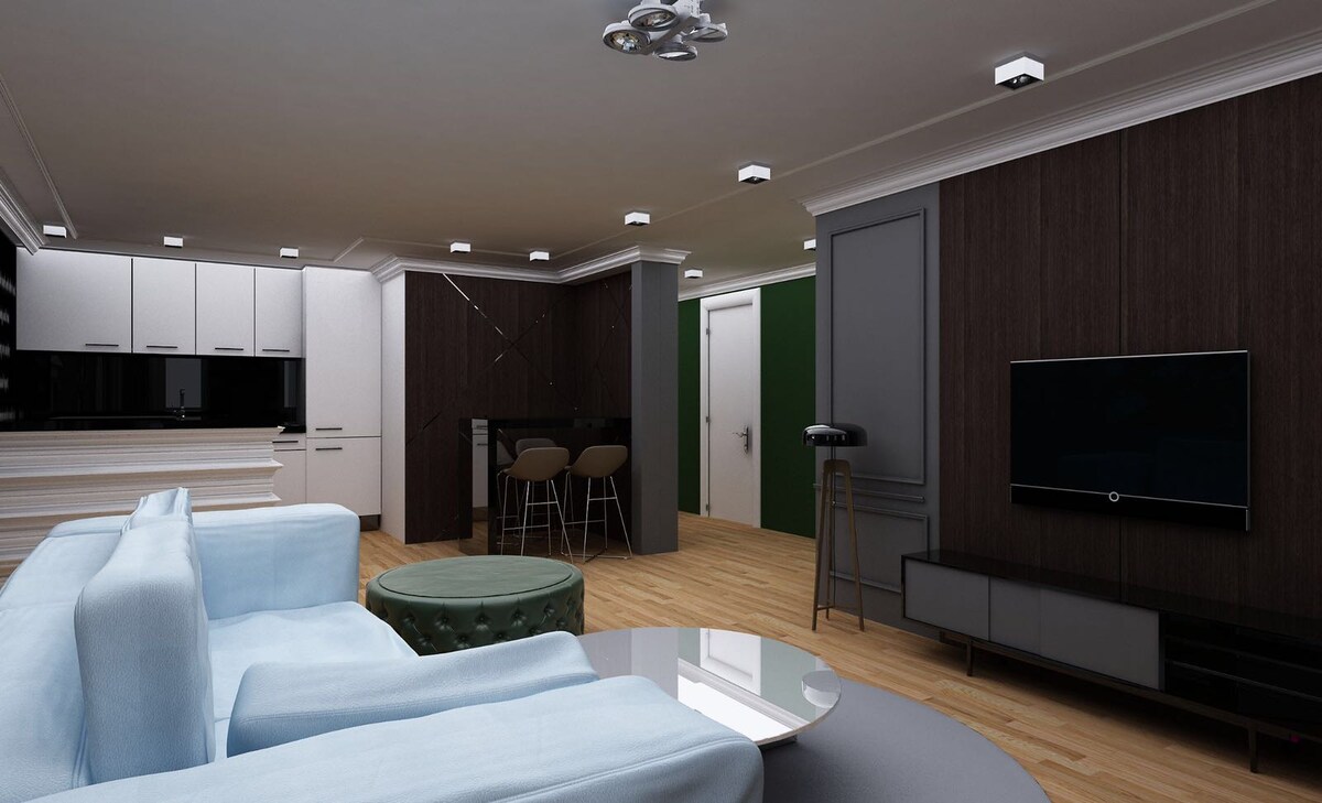 A modern living area features a light blue sectional sofa and a round coffee table, complemented by a dark green accent wall. A sleek kitchen with white cabinetry is visible in the background, and a flat-screen TV is mounted on a dark entertainment unit, enhancing the contemporary design.