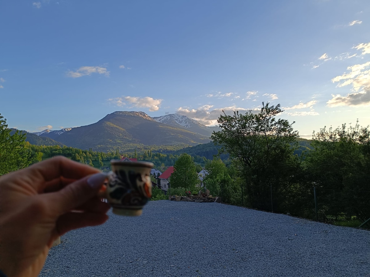 A hand holding a decorative cup is shown in the foreground, with a gravel area leading to a stunning view of mountains in the background. Lush greenery and scattered homes are visible beneath a clear blue sky with soft clouds.