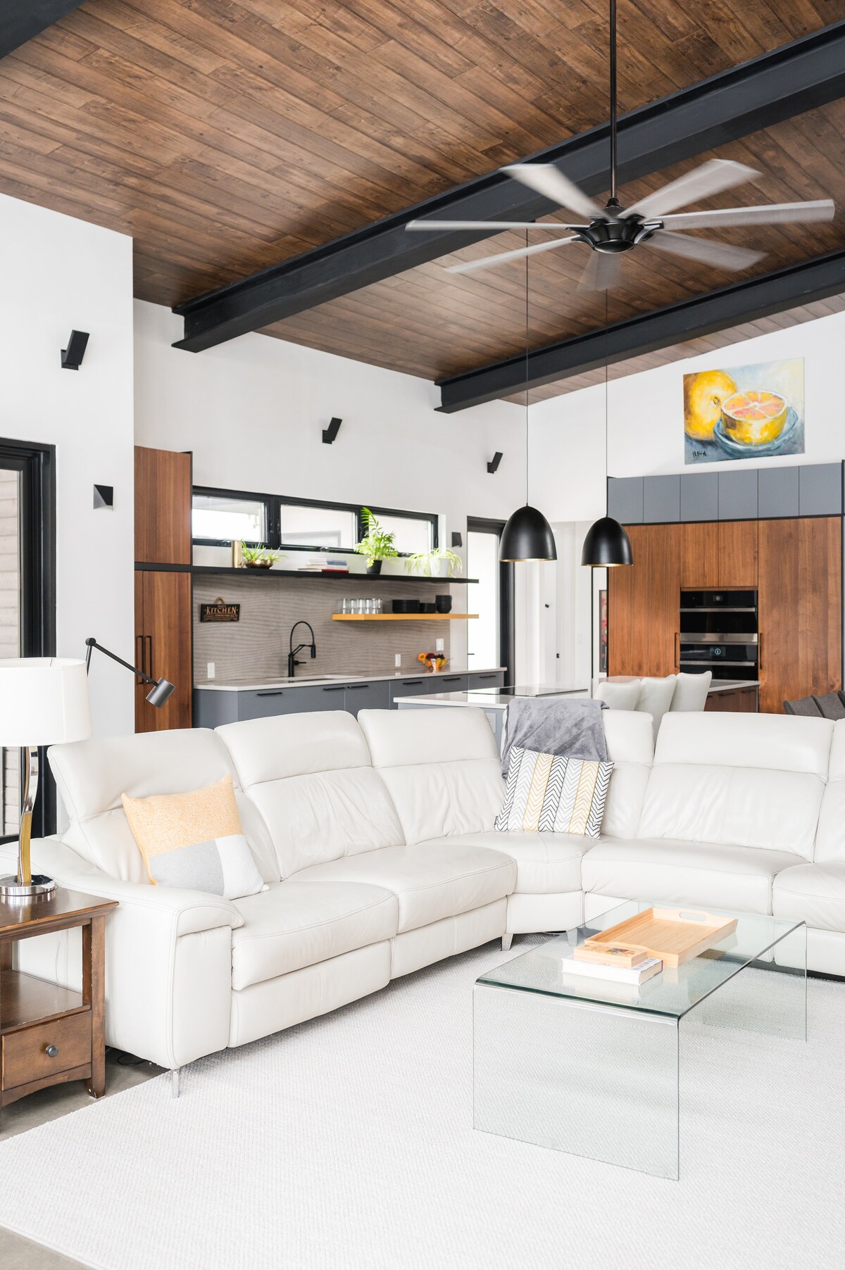 A spacious living area features a white sectional sofa arranged around a glass coffee table. Black ceiling beams contrast with the warm wooden ceiling. The open kitchen is visible in the background, showcasing modern cabinetry and a central island.