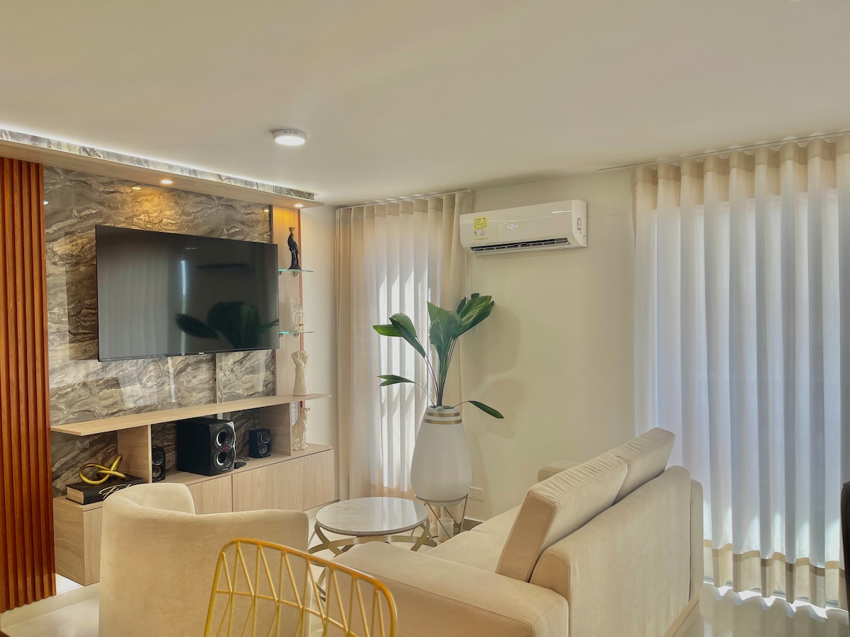 A spacious living area is designed with neutral tones, featuring a comfortable seating arrangement and a round coffee table. A flat-screen television is mounted on a textured wall, while large windows with sheer curtains allow natural light to fill the room.