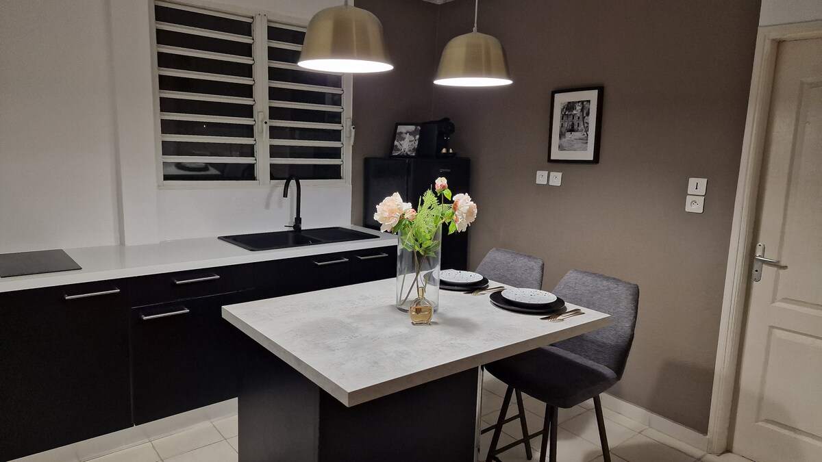 A modern kitchen area features a central island with a light-colored countertop, paired with two grey chairs. Underneath two pendant lights, a vase with fresh flowers adds a touch of color. The kitchen is equipped with a sink and sleek storage options.