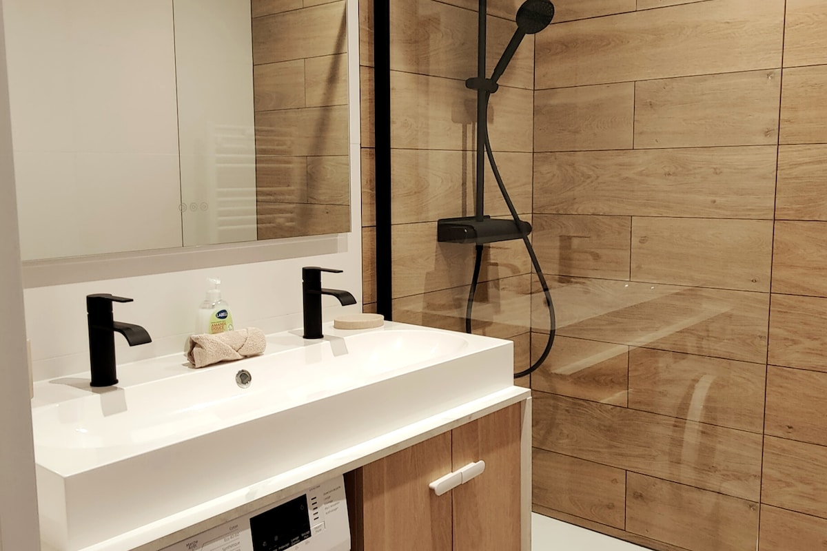 The bathroom features a modern double vanity with sleek black faucets, complemented by a large mirror. A spacious walk-in shower is enclosed with glass, showcasing minimalist fixtures. Natural wood accents on the walls contribute to a warm, contemporary aesthetic.