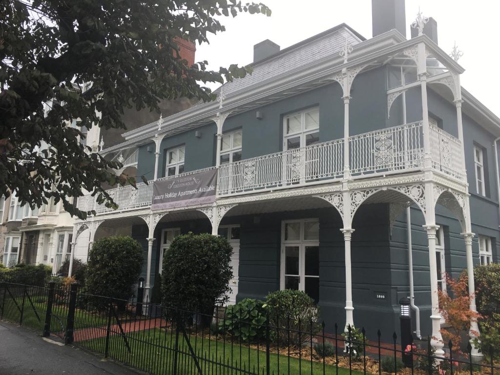 A two-story building with a striking blue facade and white decorative trim is surrounded by manicured hedges. The porch features ornate railings, and a sign indicating 'Luxury Holiday Apartments' is prominently displayed. A serene green lawn leads up to the entrance.