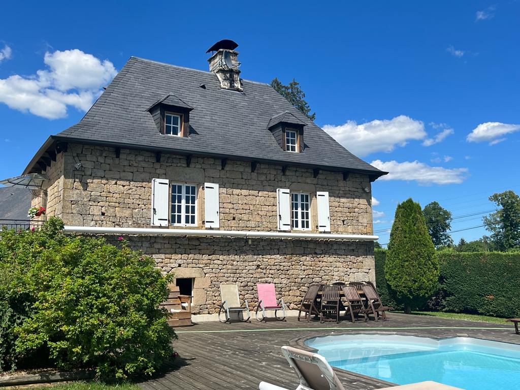 The stone house is surrounded by a well-maintained garden, featuring a swimming pool with sunbeds placed nearby. Multiple windows are present, offering natural light to the interior. The roof is completed with a chimney, and the blue sky creates a bright, inviting atmosphere.