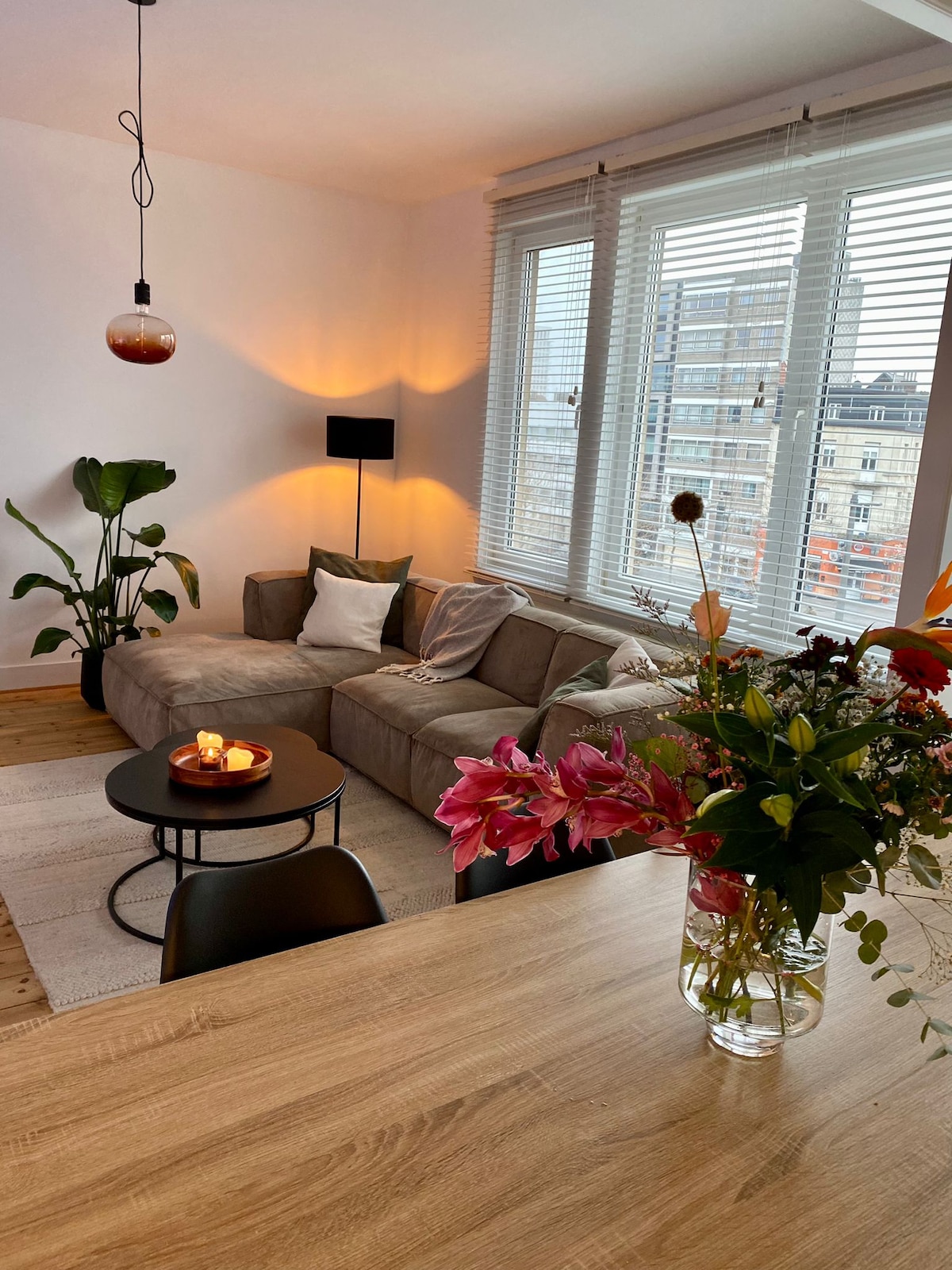 A spacious living area is highlighted by large windows allowing natural light to fill the room. A comfortable sectional sofa is complemented by a small round coffee table and decorative plants. A dining table is adorned with a vibrant flower arrangement, enhancing the inviting atmosphere.