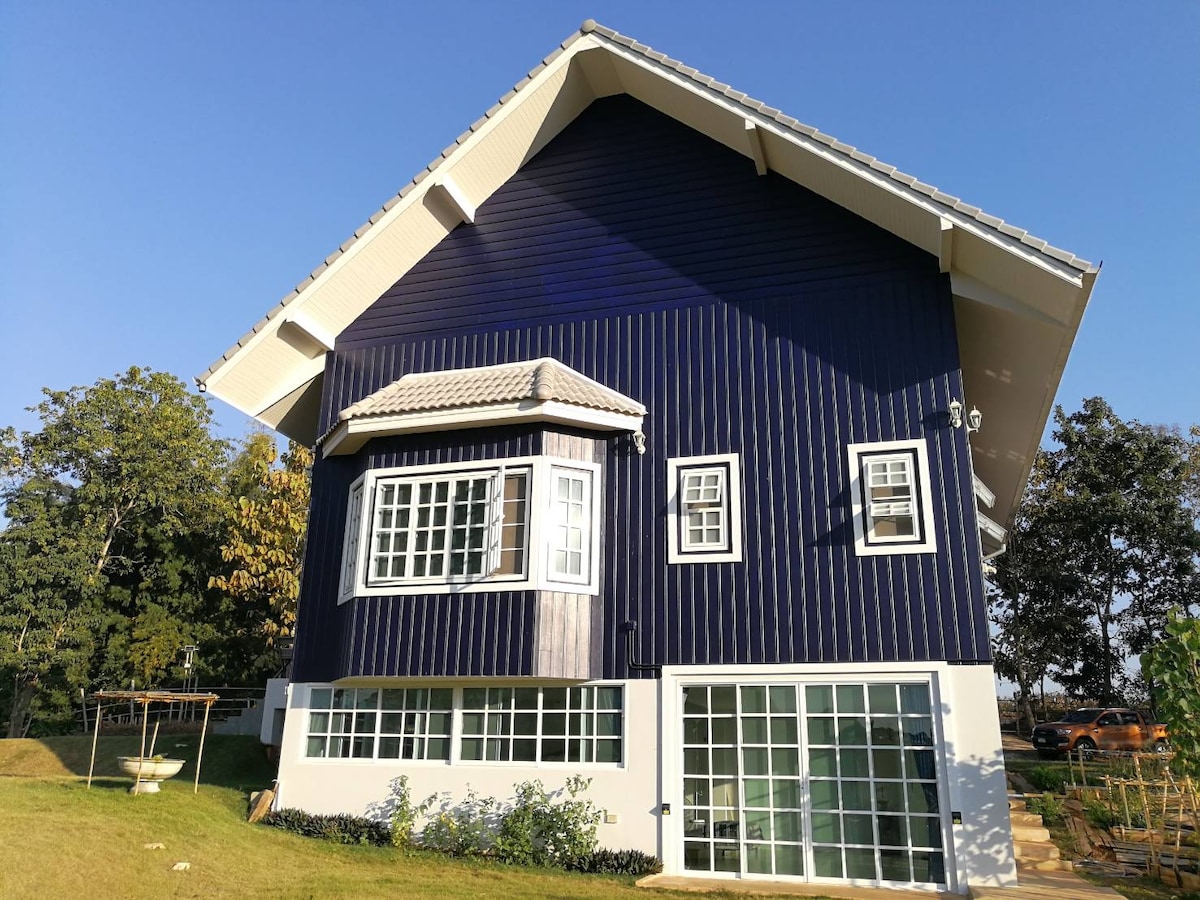 A two-story, blue exterior house features large windows, allowing natural light to fill the interior. The lower level showcases multiple glass doors that provide a view of the surrounding greenery. The sloped roof contributes to the home's modern, charming design.