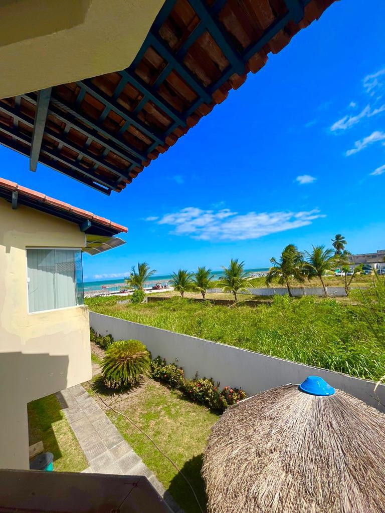A view from the balcony showcases a vibrant blue sky and lush greenery, with palms lining the coastline in the distance. A thatched gazebo is positioned in the foreground, providing a shaded area in the outdoor space.