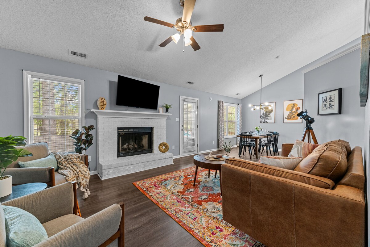 A spacious living area features a wooden ceiling fan and a cozy seating arrangement, including a brown couch and light-colored armchairs. Natural light filters through the windows, illuminating the room's soft blue walls. A TV is mounted above a white fireplace, while a dining space is seen in the background.