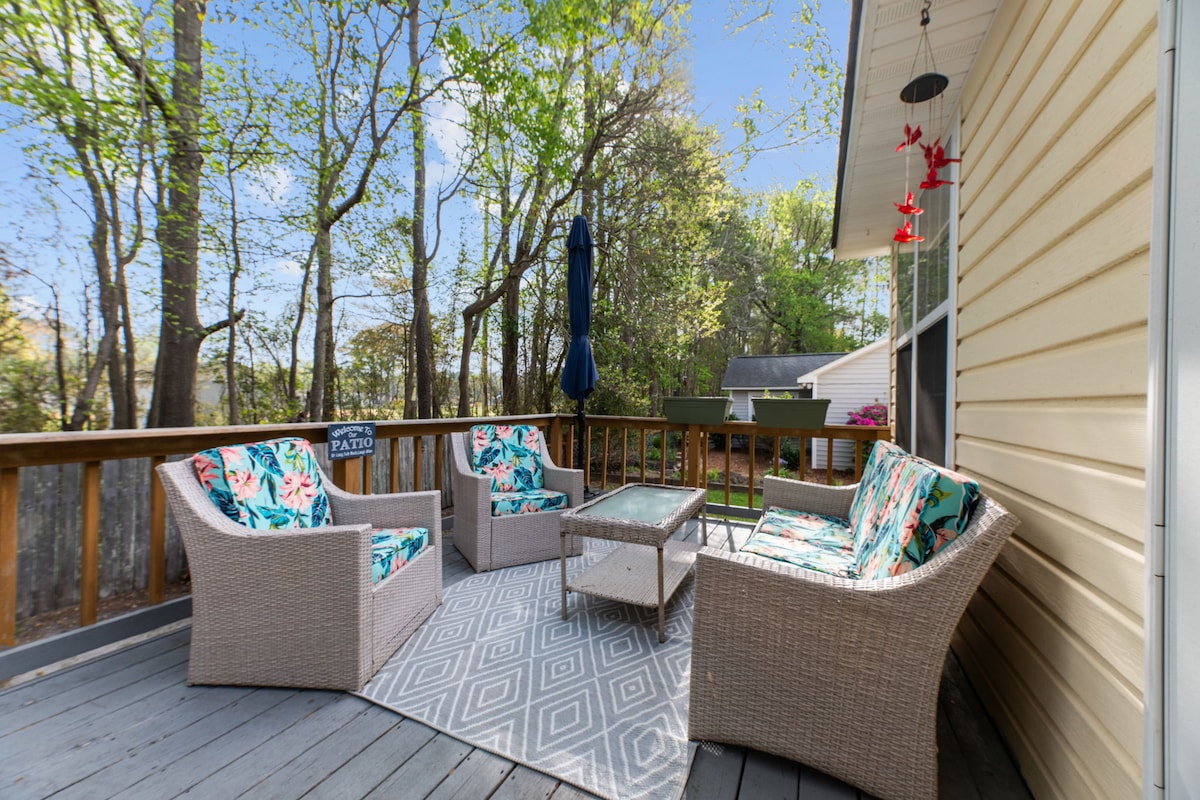 An outdoor seating area is featured on a shaded deck, showcasing four wicker chairs adorned with colorful floral cushions. A glass-top table sits at the center, complemented by a patterned rug. Lush greenery and trees provide a natural backdrop to the space.