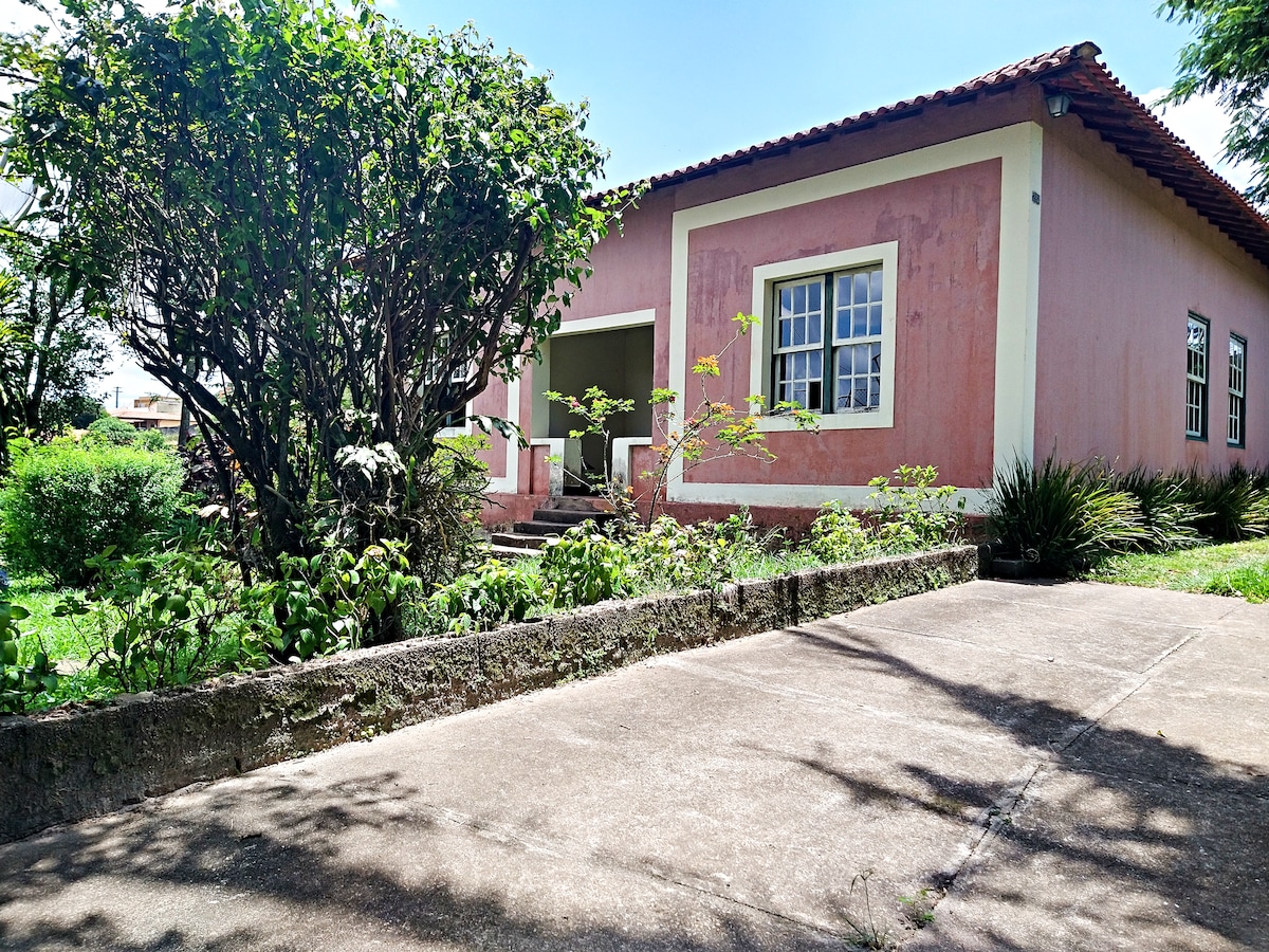 A rustic house with a pink exterior is framed by a lush green garden. A walkway leads to the entrance, bordered by ornamental plants and trees. Large windows allow natural light to fill the interior spaces.