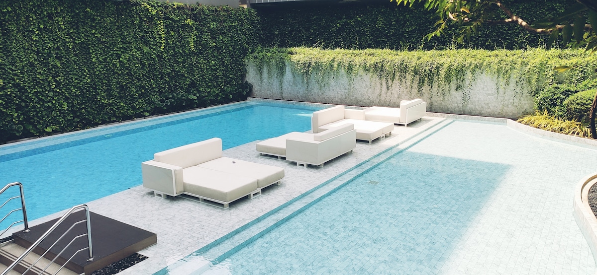 A sunken pool area is surrounded by lush greenery, featuring white lounge sofas positioned partially in the water. The pool's clear blue surface reflects the natural surroundings, creating a serene atmosphere. A poolside deck with a staircase provides access to the water.