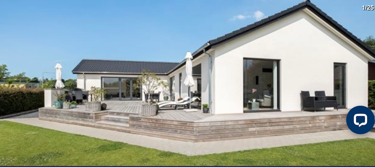 The exterior of the home showcases a spacious wooden terrace, furnished with lounge chairs and shaded by umbrellas. Lush green grass surrounds the property, while expansive glass doors provide a view of the light-filled interior, enhancing the welcoming atmosphere of the residence.