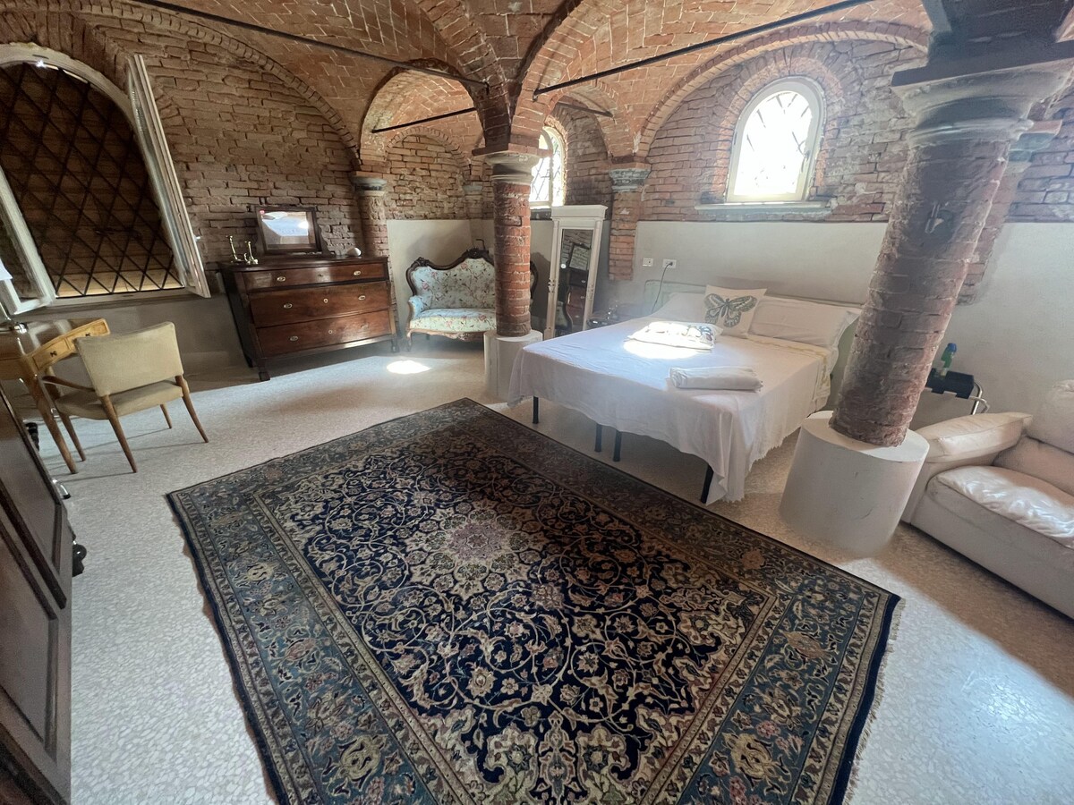 A spacious room features exposed brick arches and large windows. A wooden chest of drawers and an ornate sofa are present alongside a neatly made bed. A decorative rug covers the floor, adding warmth to the environment.