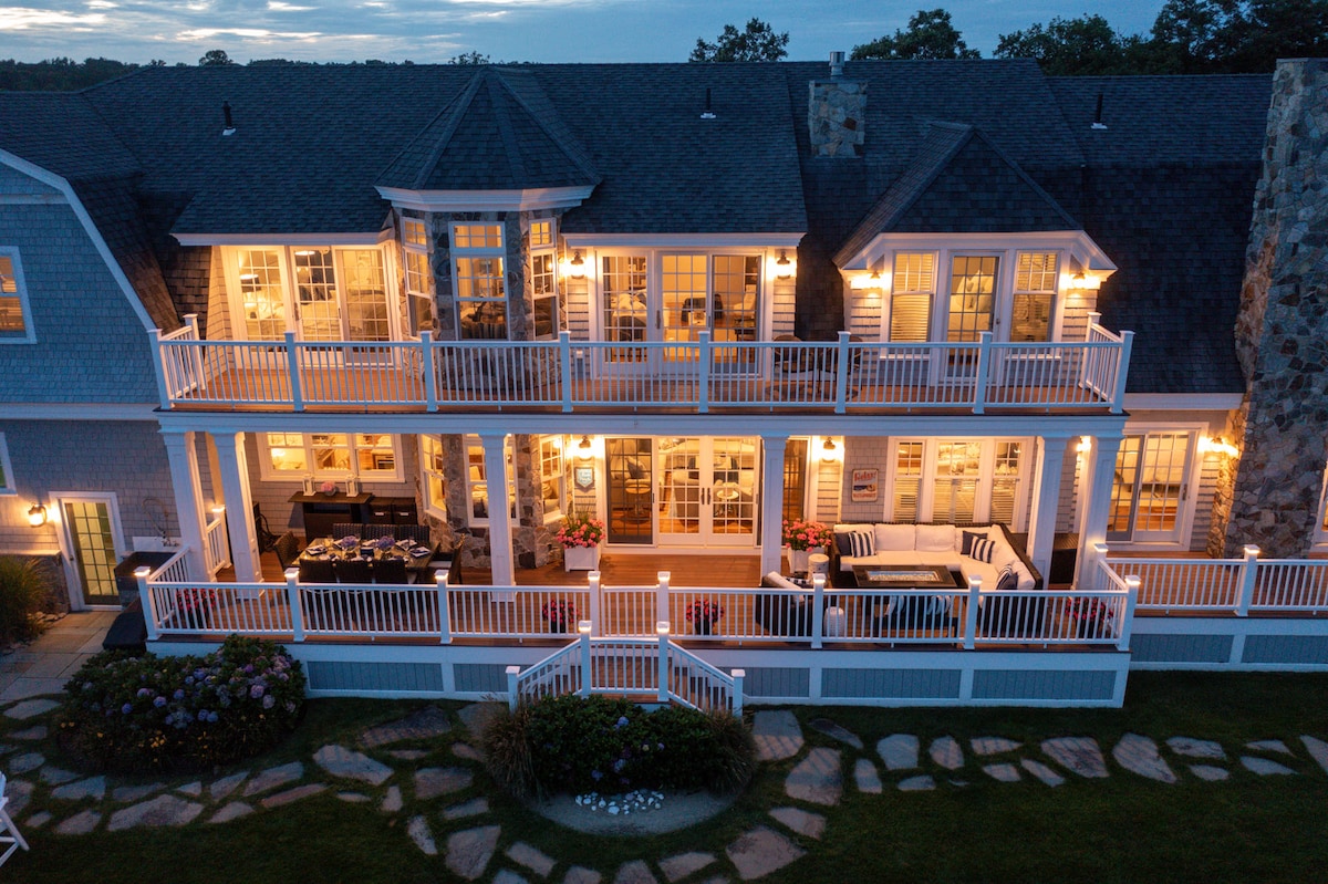 The two-story exterior of the house is showcased at dusk, illuminated by warm lighting. A spacious deck features seating areas for relaxation, with sliding glass doors leading to indoor living spaces. A well-maintained pathway and lush landscaping enhance the inviting appearance of the home.