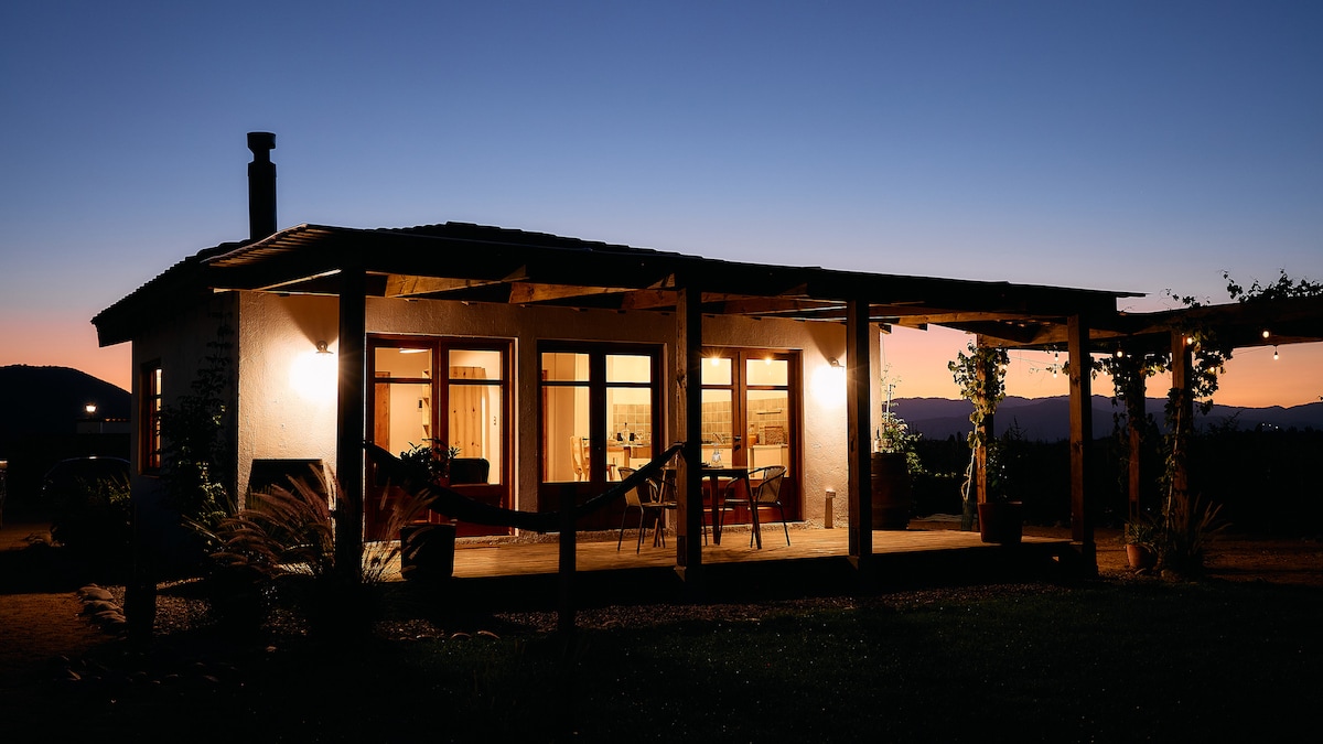 The exterior of a rustic chalet is shown at dusk, featuring a porch with wooden beams. Soft lighting highlights large windows, revealing the warm interior. A hammock hangs gently beneath the structure, with surrounding greenery and distant hills visible in the background.