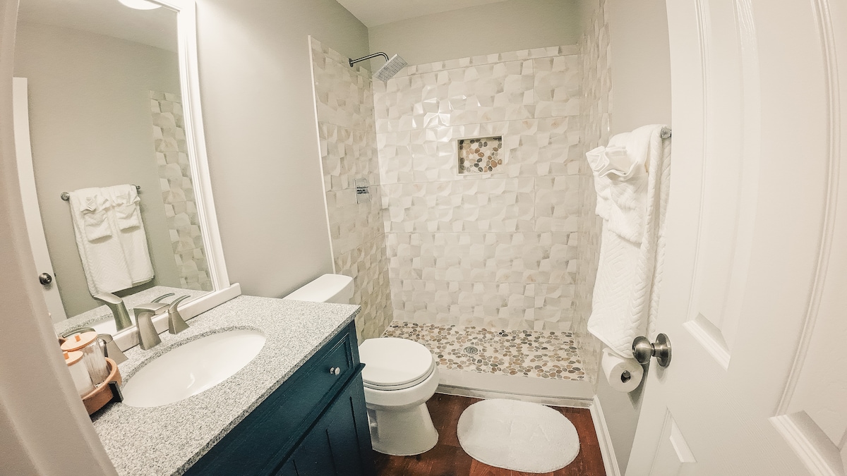 A modern bathroom features a tiled shower with a decorative niche and an elegant countertop with a sink. Soft white towels are neatly arranged on a rack. A round bath mat is positioned in front of the shower, and natural light brightens the space.