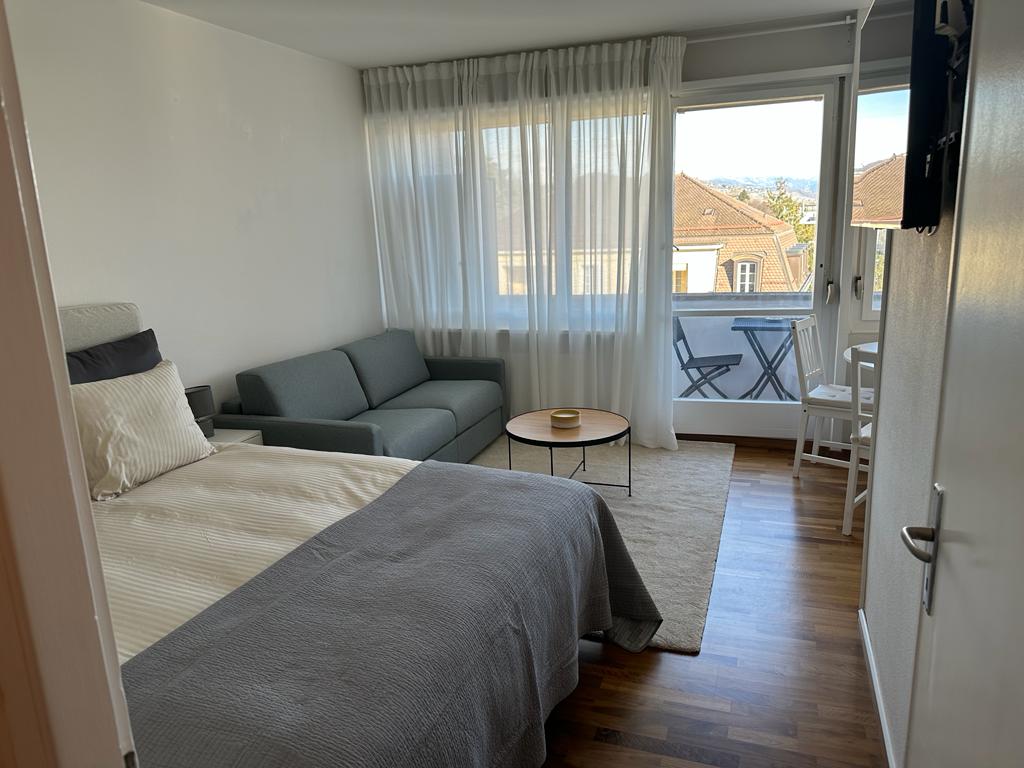 A bright studio space features a spacious bed, a cozy sofa, and a small round table set beside large windows. Sheer curtains diffuse natural light, creating a serene ambiance. Outdoor seating is visible on the balcony, enhancing the connection to the outside.