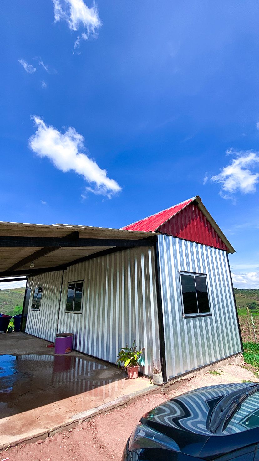 A modern metal-clad structure is captured under a bright blue sky, featuring a red roof and large windows that allow natural light in. A small potted plant is positioned near the entrance, adding a touch of greenery to the exterior space.